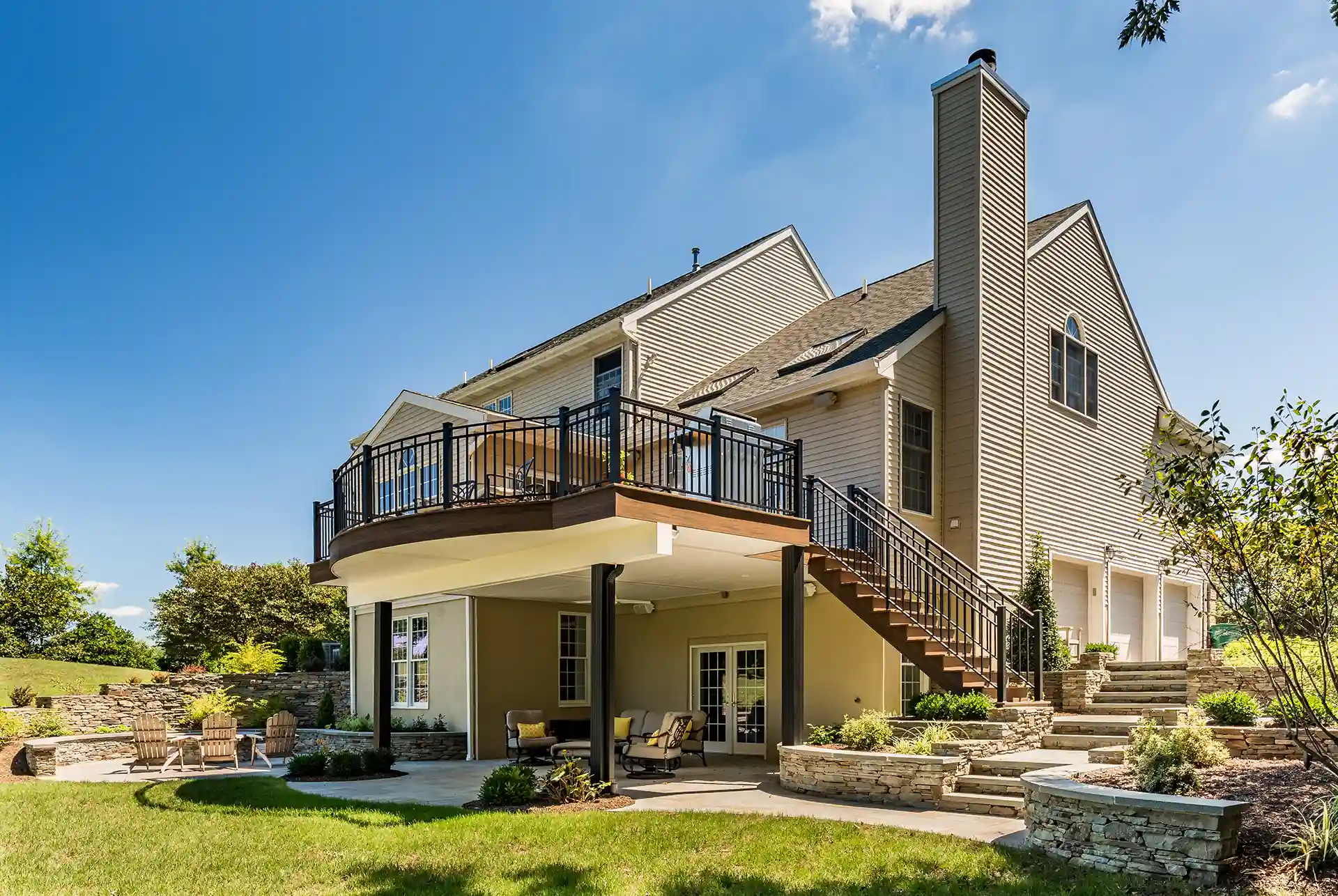 Covered stone patio beneath elevated deck with outdoor seating, stairs, and tiered garden steps.
