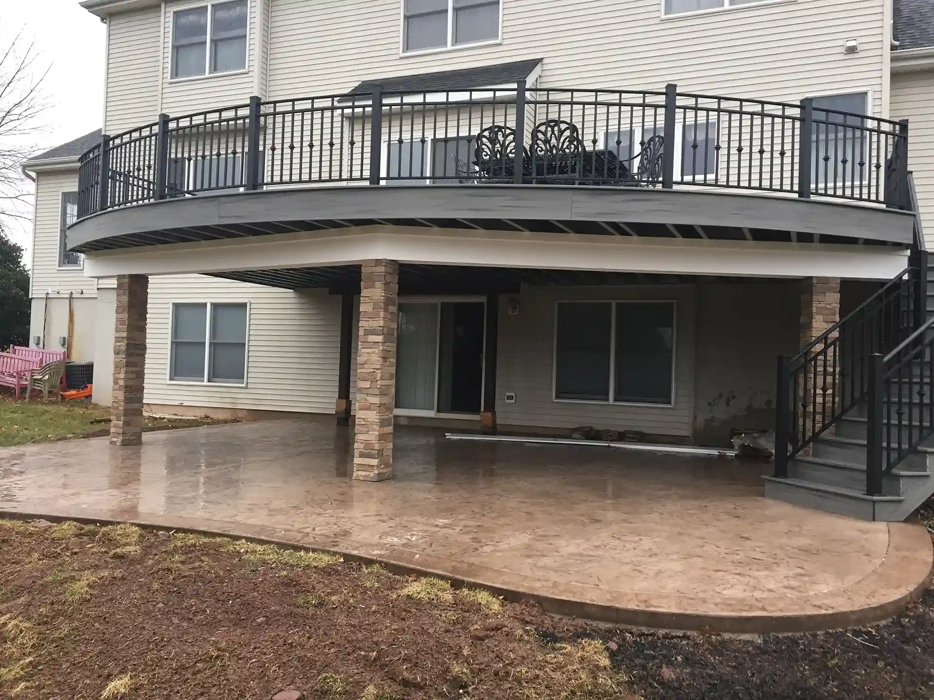 Stamped concrete patio beneath curved deck with stone columns and black metal railing above.
