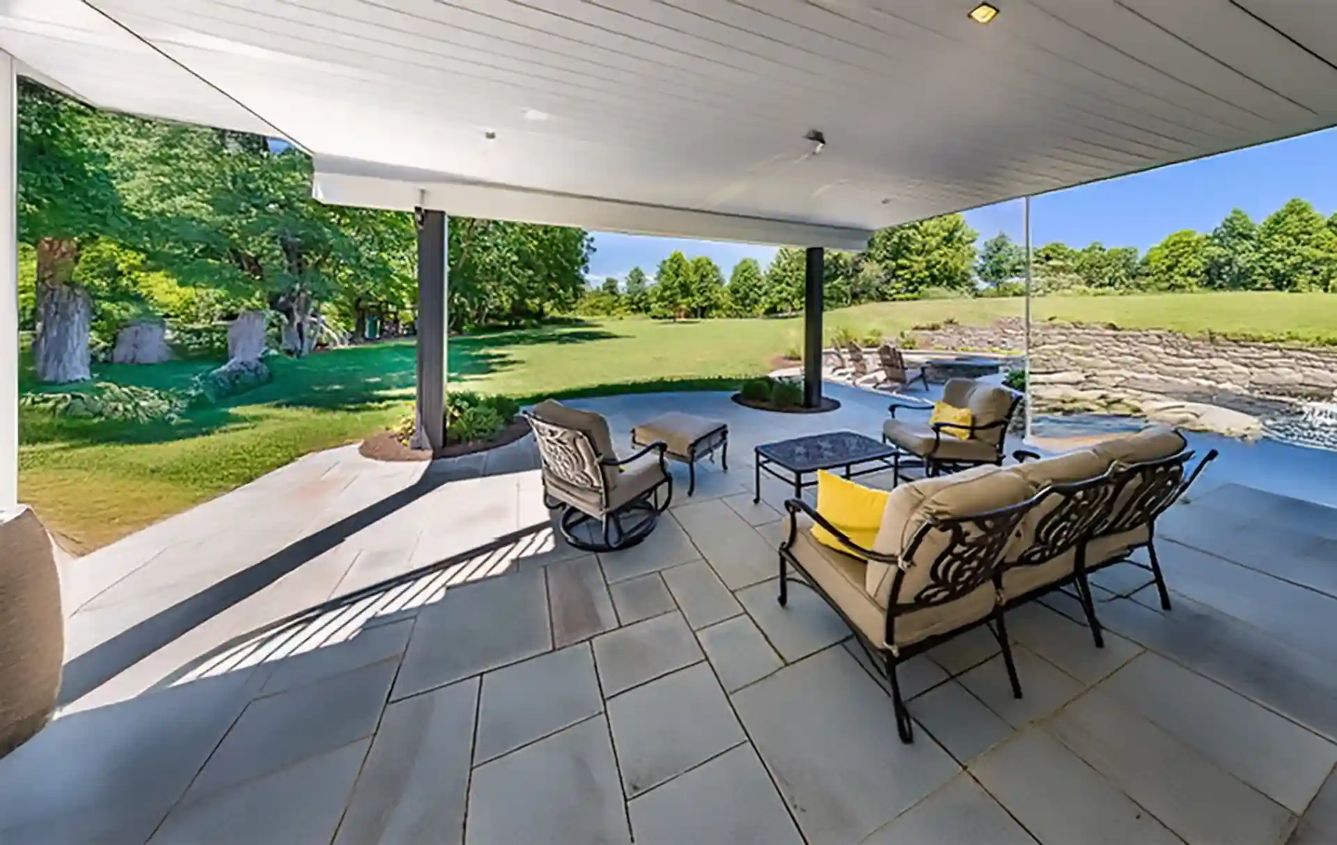 Under-deck patio with stone flooring, cushioned metal chairs, and open view of grassy yard.
