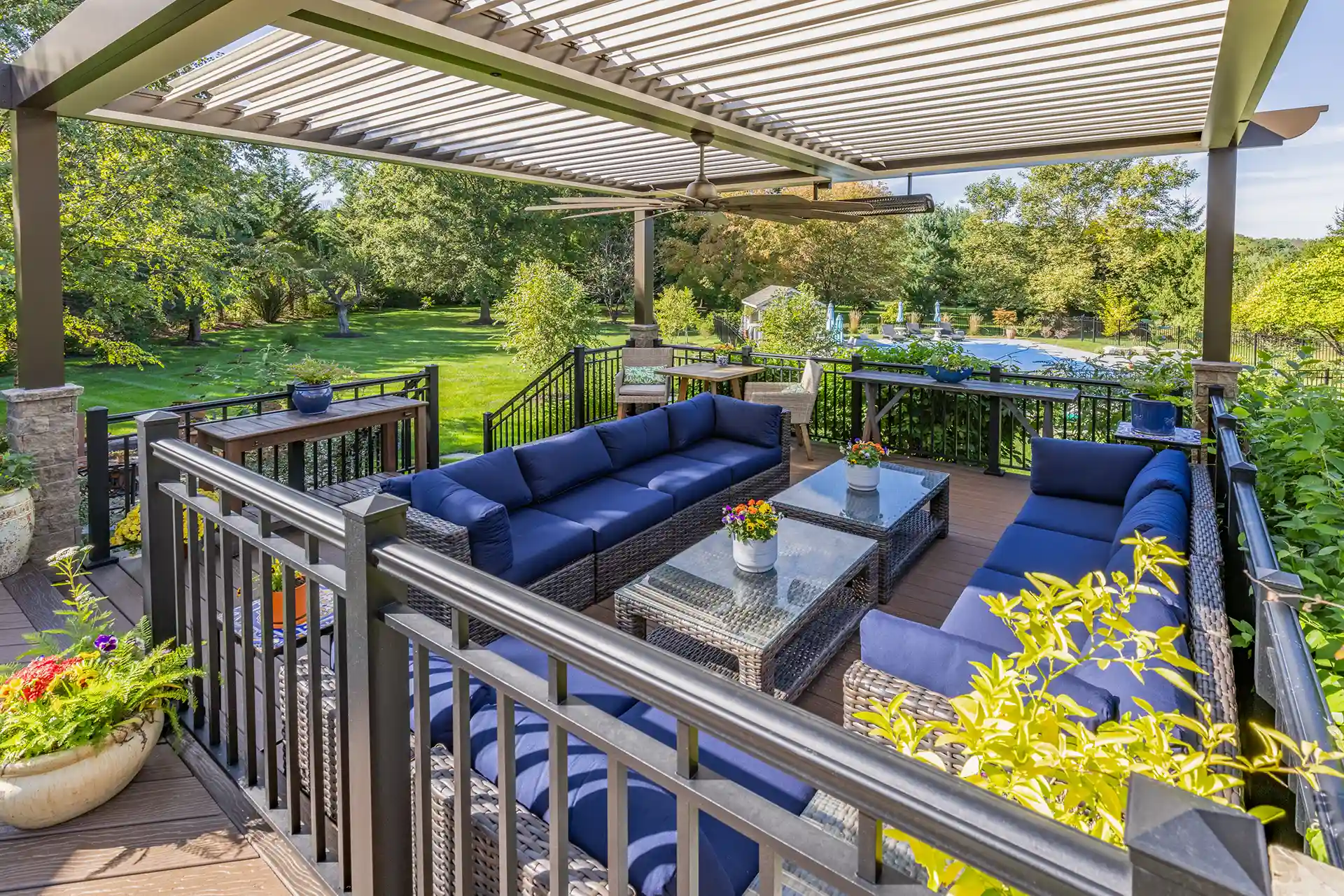Phot of a louvered roof pergola providing cover for an elaborate outdoor living room on a deck with deck railing and deck steps.